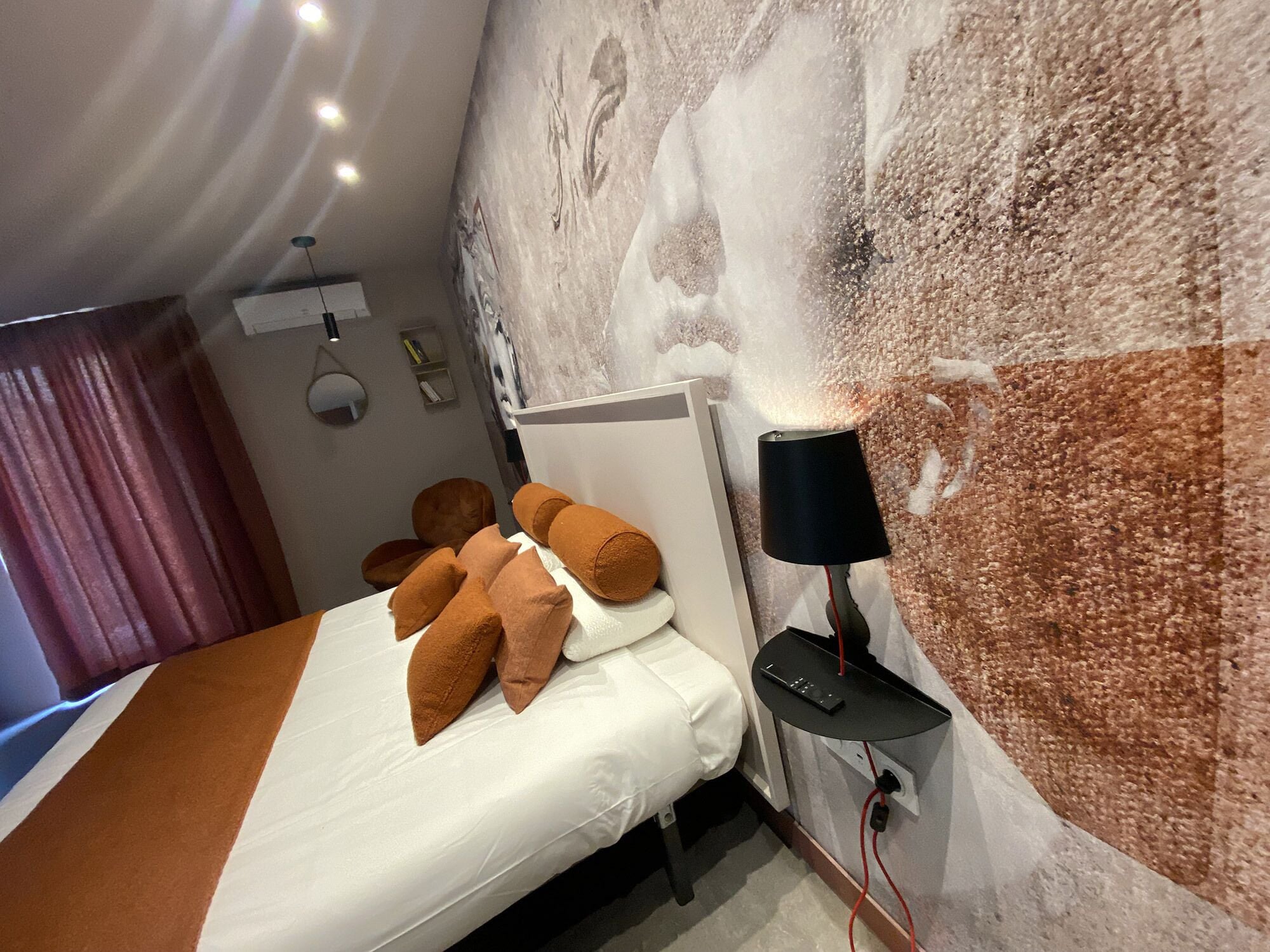 Un llit ben fet amb naxelles marrons i blanques, una làmpada negra sobre una taula de nit i una paret amb un mural abstracte gran.