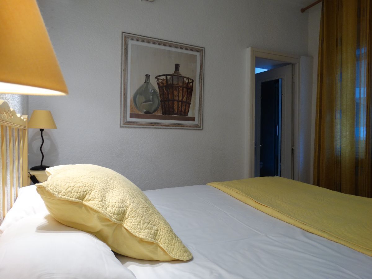 A neatly made bed with a yellow blanket and pillow, a framed picture on the wall, and a lamp on a bedside table.