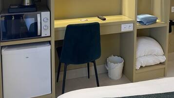 Habitación de hotel compacta con kitchenette, un televisor de pantalla plana montado en la pared, un escritorio con silla y una cama con almohada blanca.