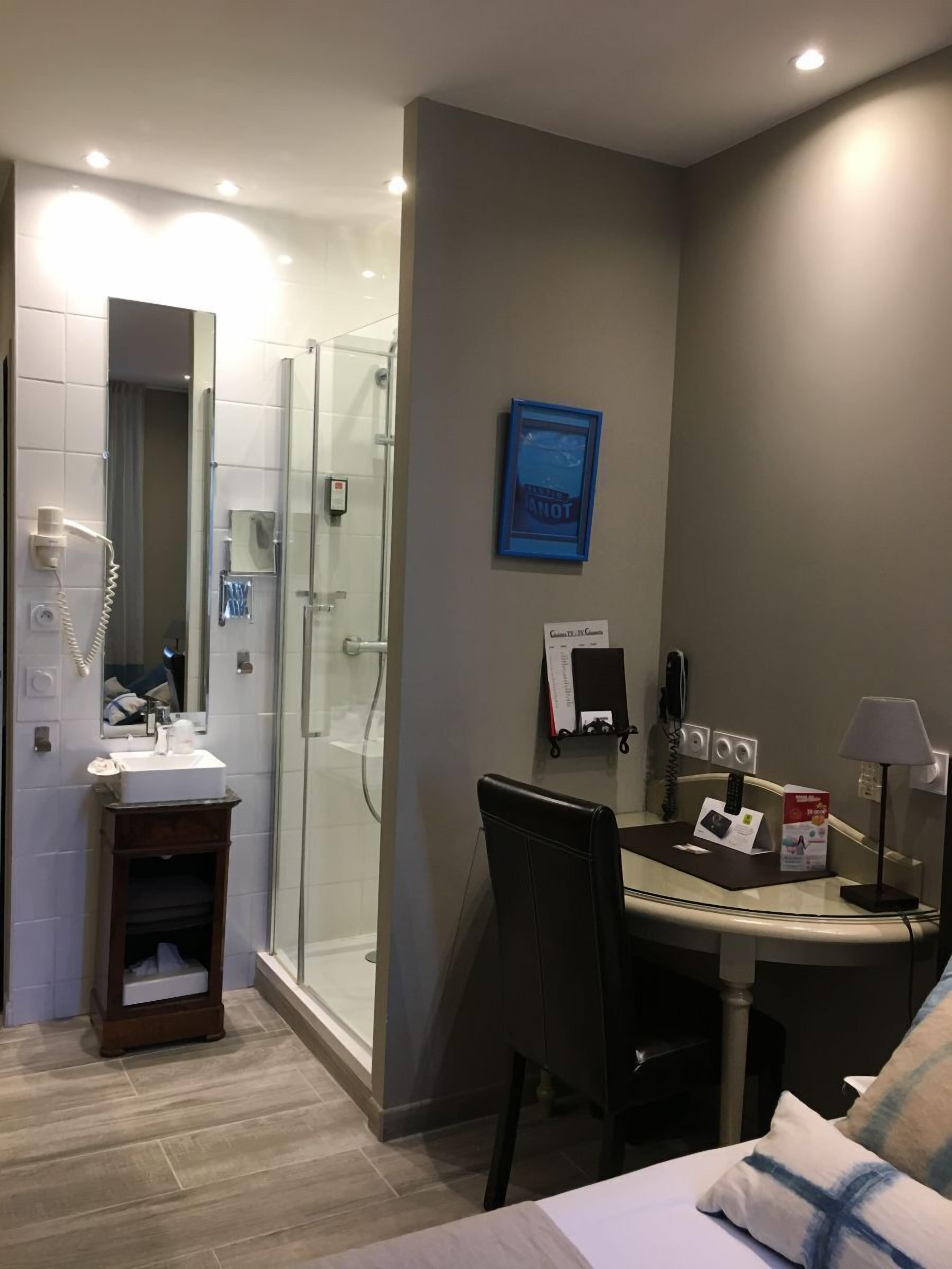 A modern bathroom with a glass shower enclosure, a sink with a mirror, and a vanity. There is a desk with a chair, a lamp, and a telephone. A framed picture is on the wall.