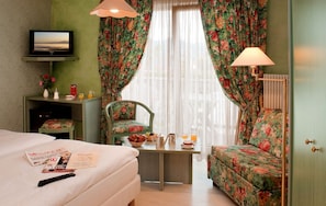 Una habitación de hotel con cama, escritorio con silla, televisor y ventana con cortinas.