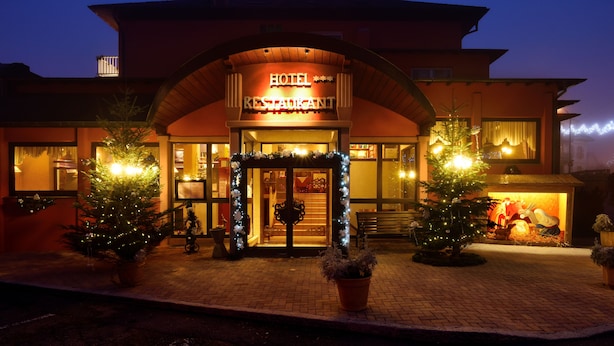 L’entrée d’un hôtel, décorée d’un sapin de Noël et de guirlandes lumineuses, avec une enseigne indiquant « HÔTEL-RESTAURANT », et un chemin pavé menant à l’entrée.