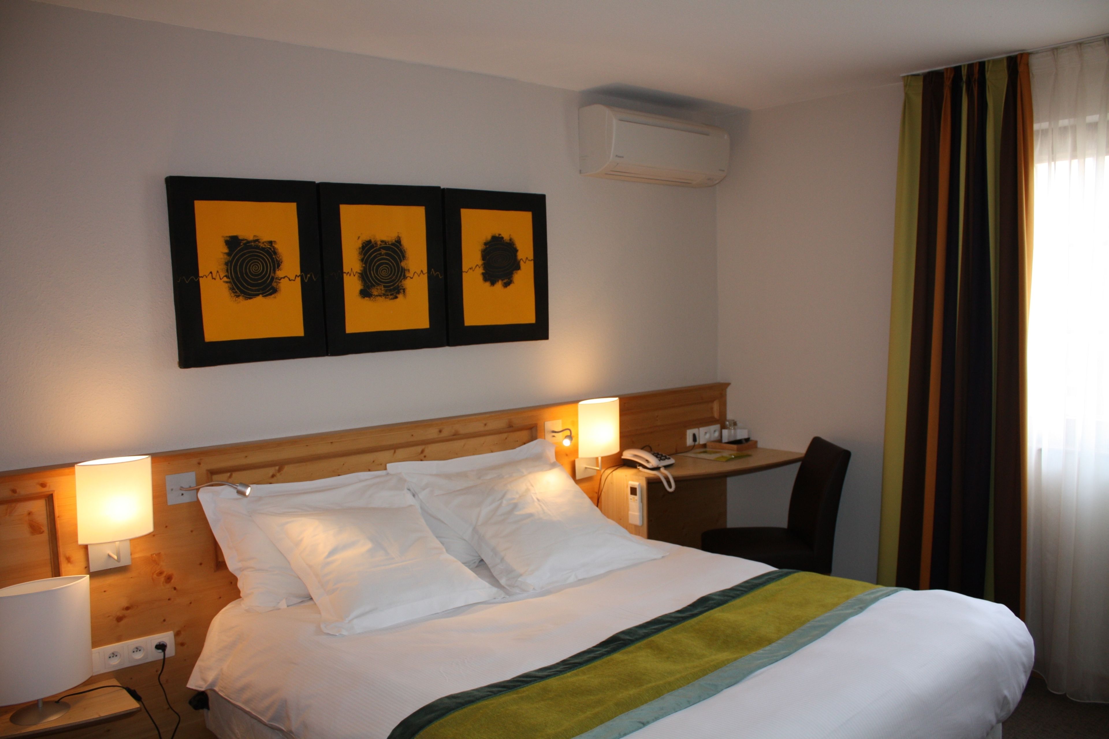 Habitación de hotel con cabecera de madera, cama con ropa de cama blanca, dos lámparas de noche, un escritorio con teléfono y tres cuadros en la pared.