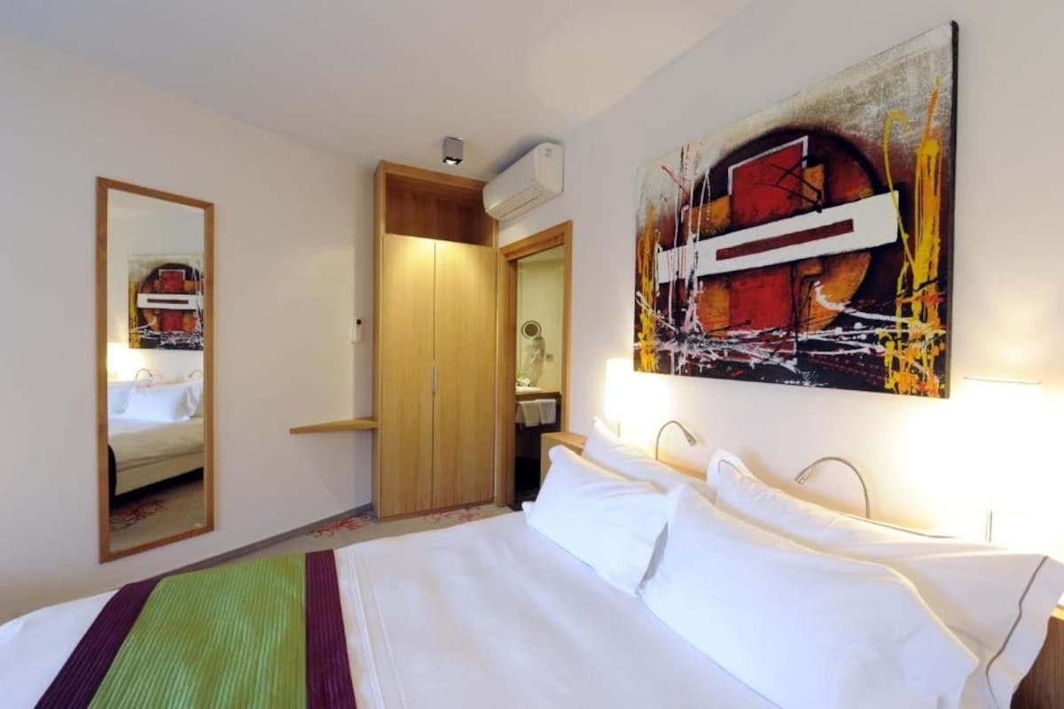 A hotel room with a bed, a mirror, a wardrobe, and a painting on the wall.