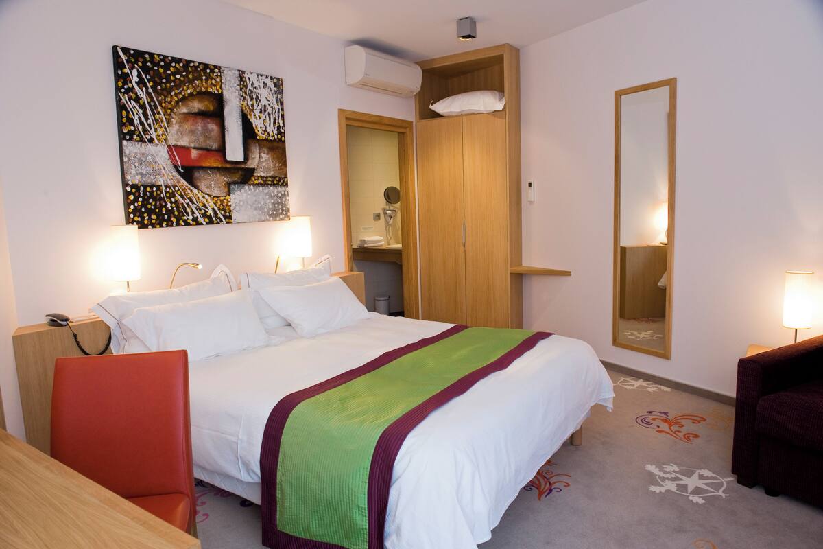 A hotel room with a bed, a desk, a chair, a wardrobe, a mirror, and a painting on the wall.