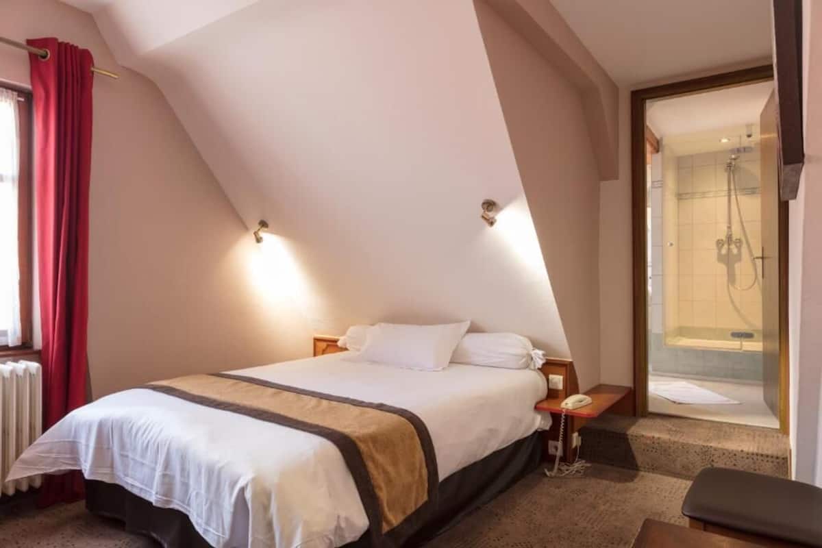 A hotel room with a bed, a bedside telephone, a shower area, and a window with red curtains.