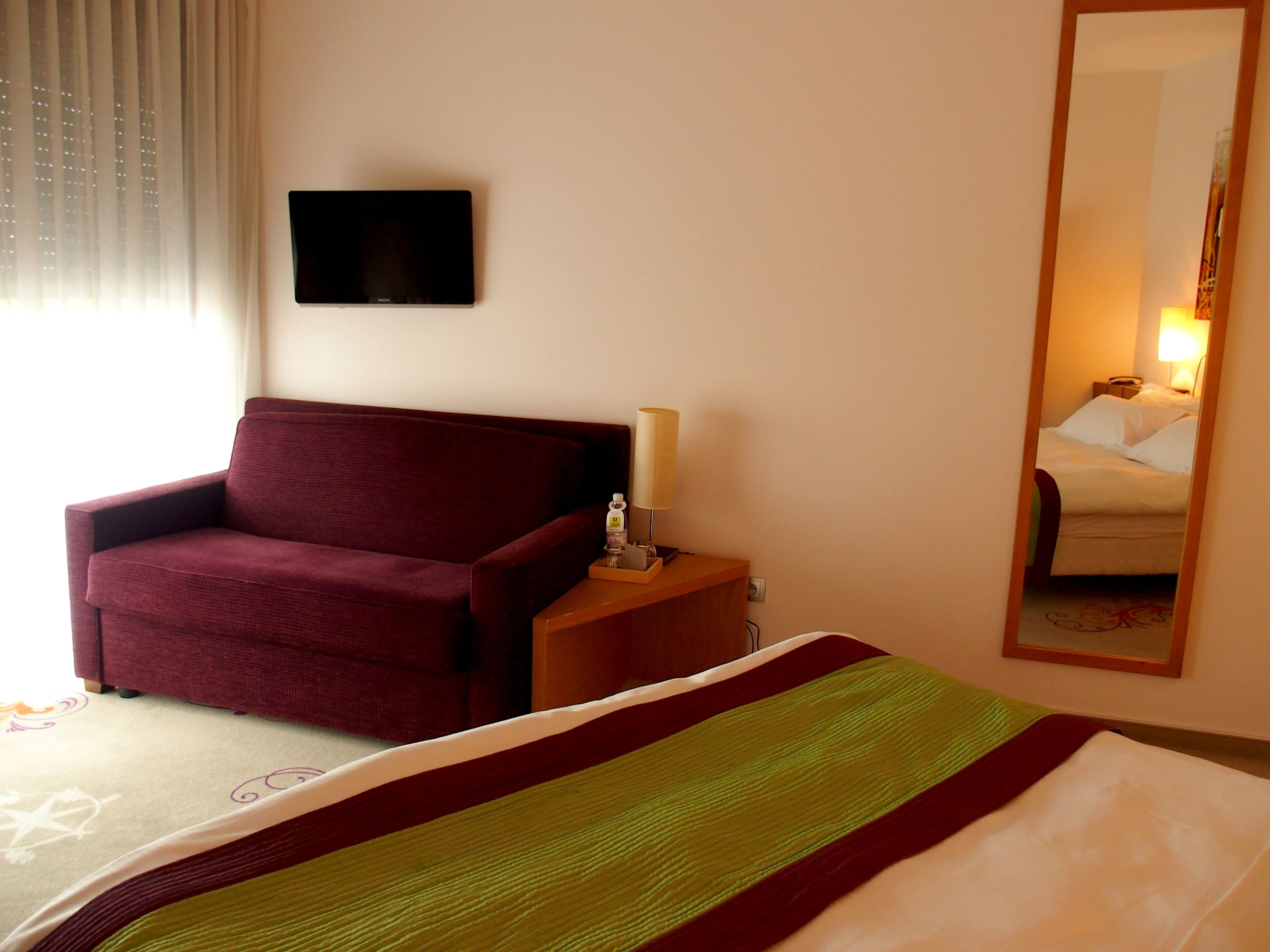 A hotel room with a bed, a sofa, a bedside table with a lamp, and a TV on the wall.