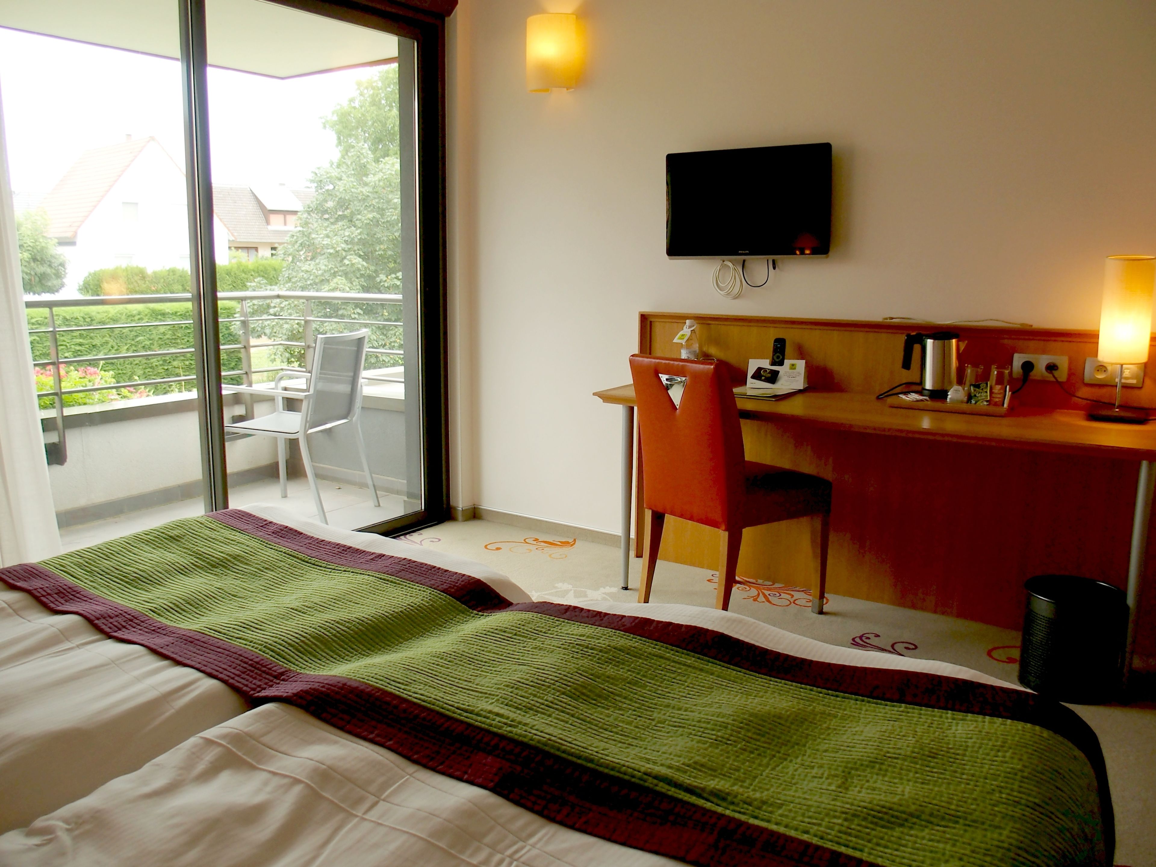 A hotel room with a bed, a desk, a chair, a TV, and a balcony with a chair.