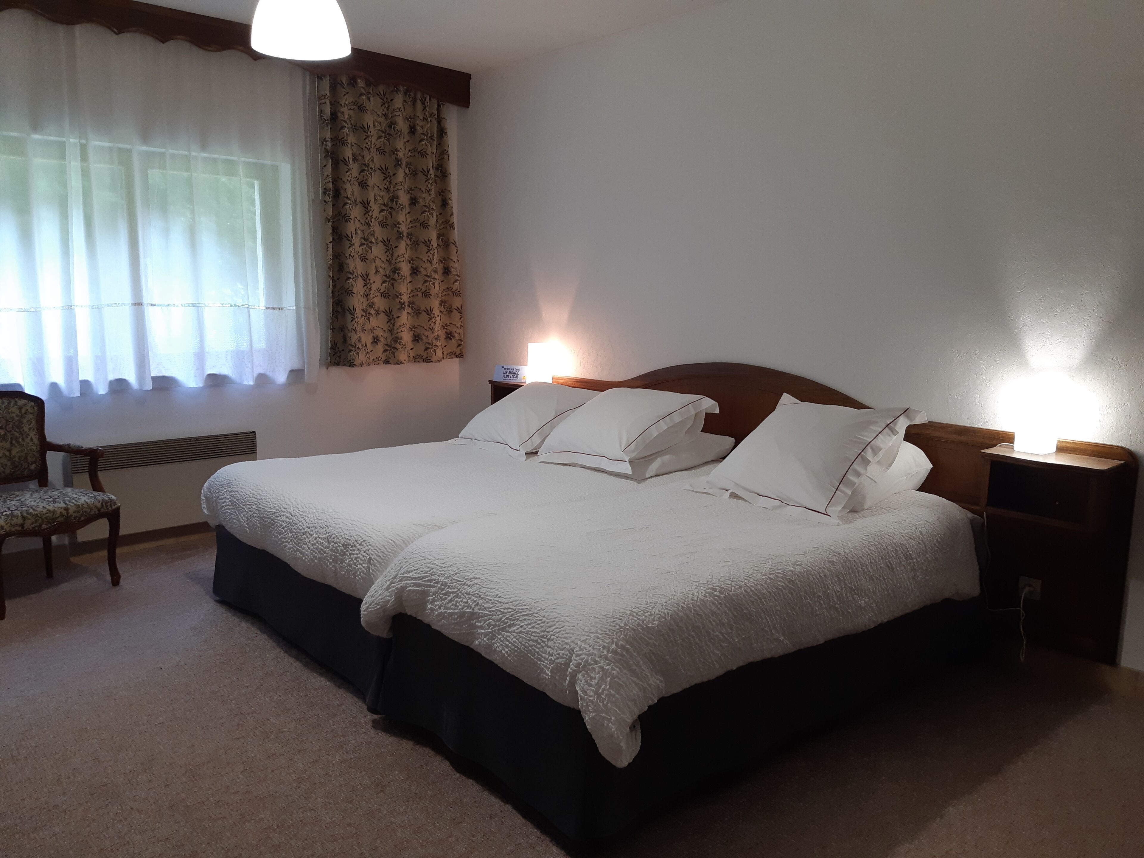Een netjes ingerichte hotelkamer met een groot bed, twee nachtkastjes met lampen, een stoel en een raam met dunne gordijnen.