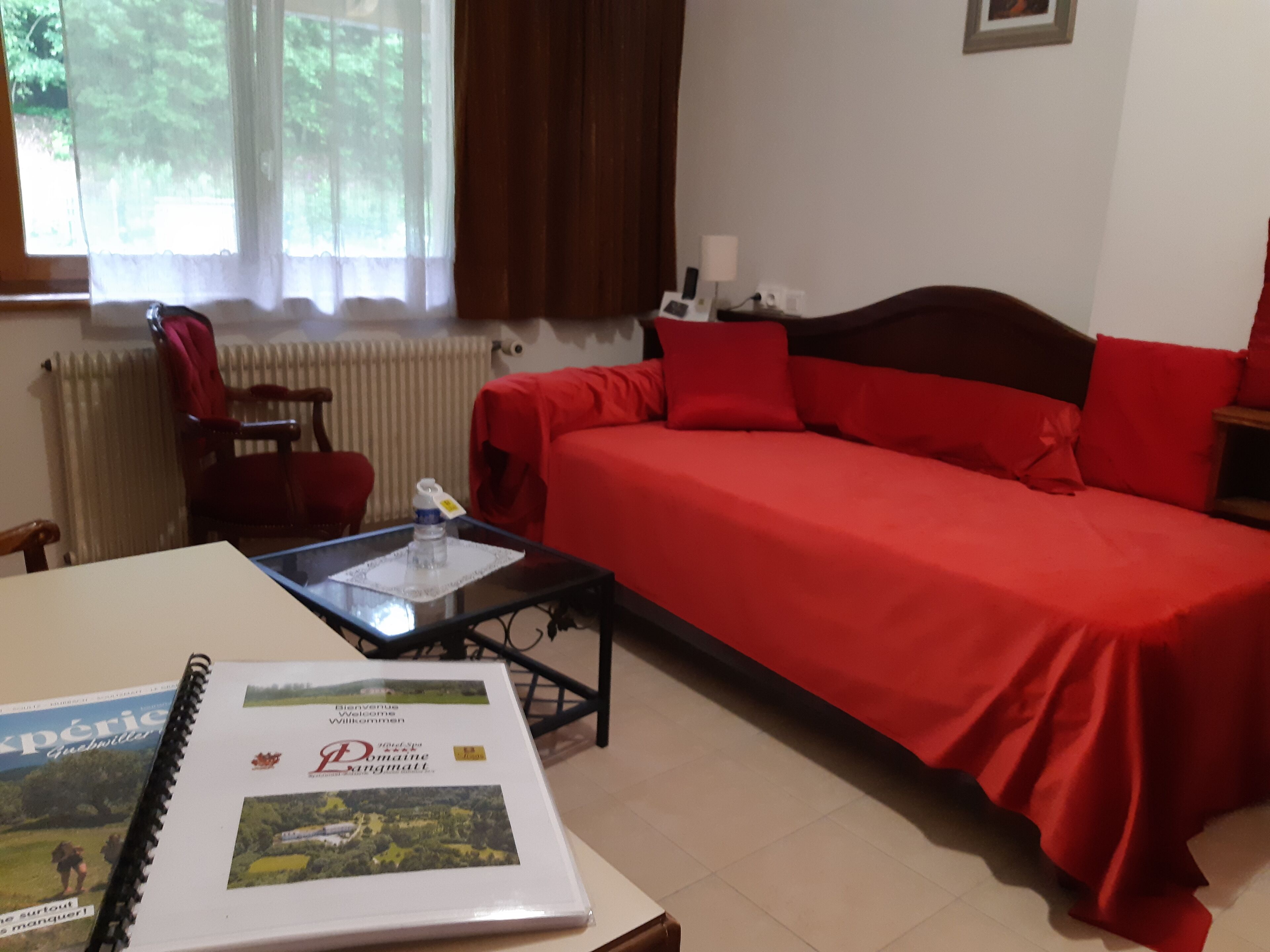 A room with a bed, a table with a book, a chair, and a window with curtains.