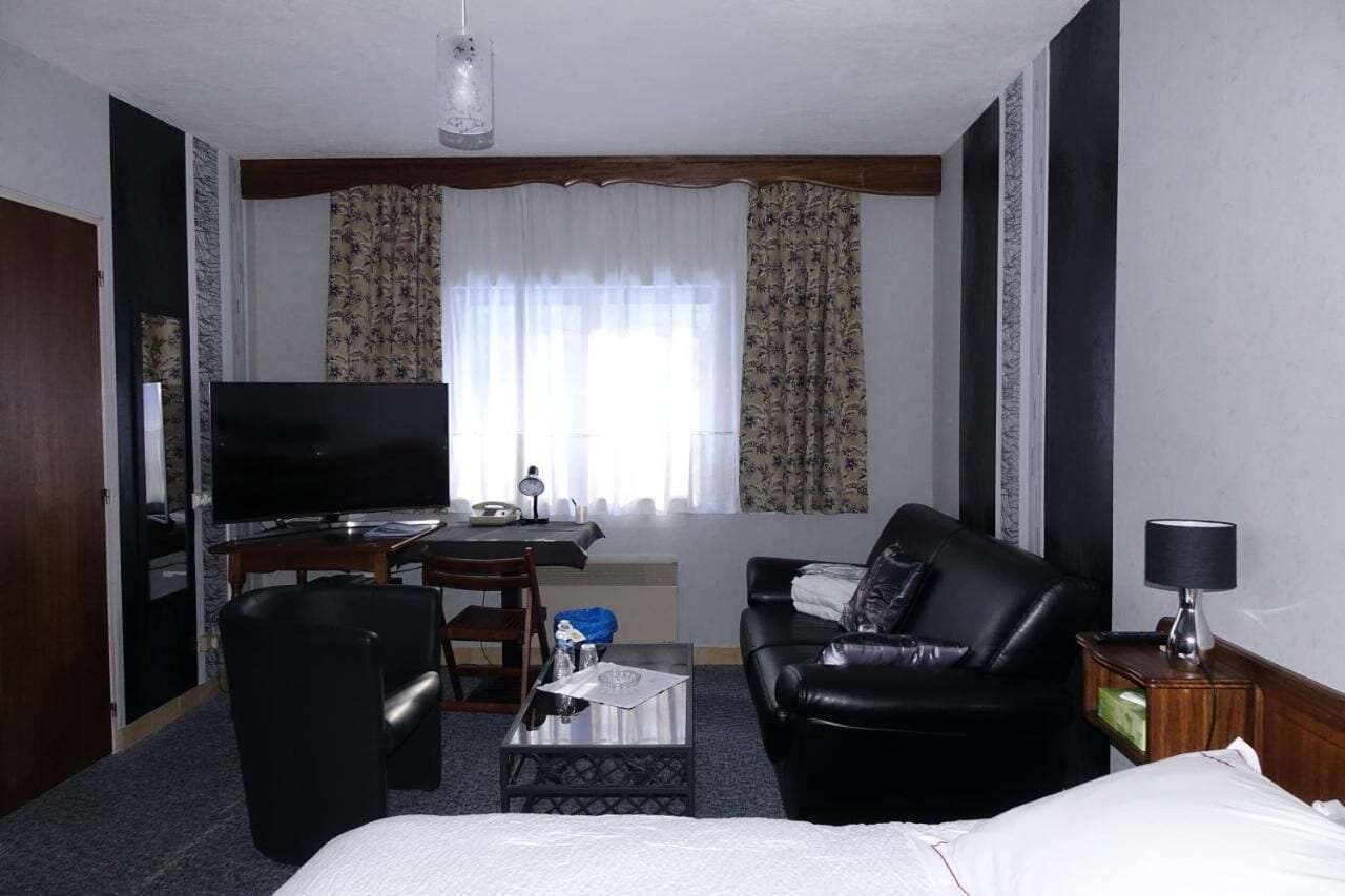 A hotel room with a bed, a black leather sofa, a television, a desk with a lamp, and a window with curtains.
