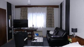 A hotel room with a bed, a black leather sofa, a television, a desk with a lamp, and a window with curtains.