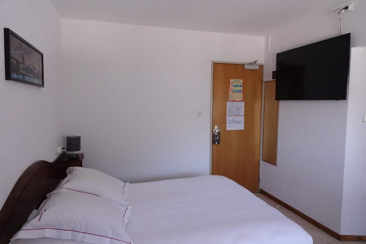 A hotel room with a bed, a wooden door, a mounted TV, and a small table with a lamp.