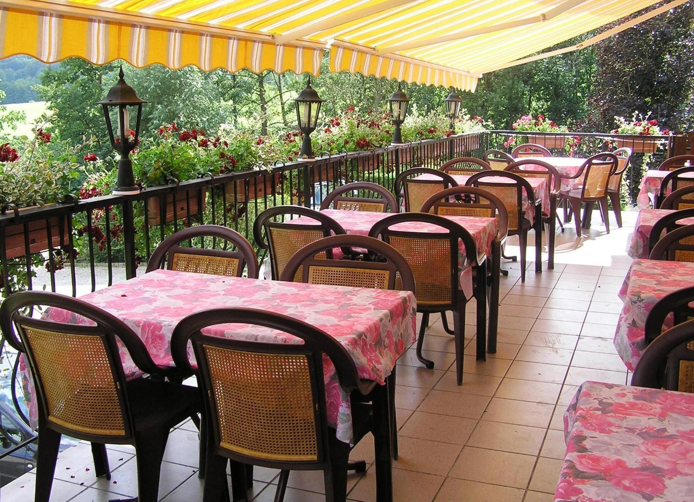 An outdoor patio with tables and chairs, covered by a yellow canopy, and surrounded by greenery and flowers.