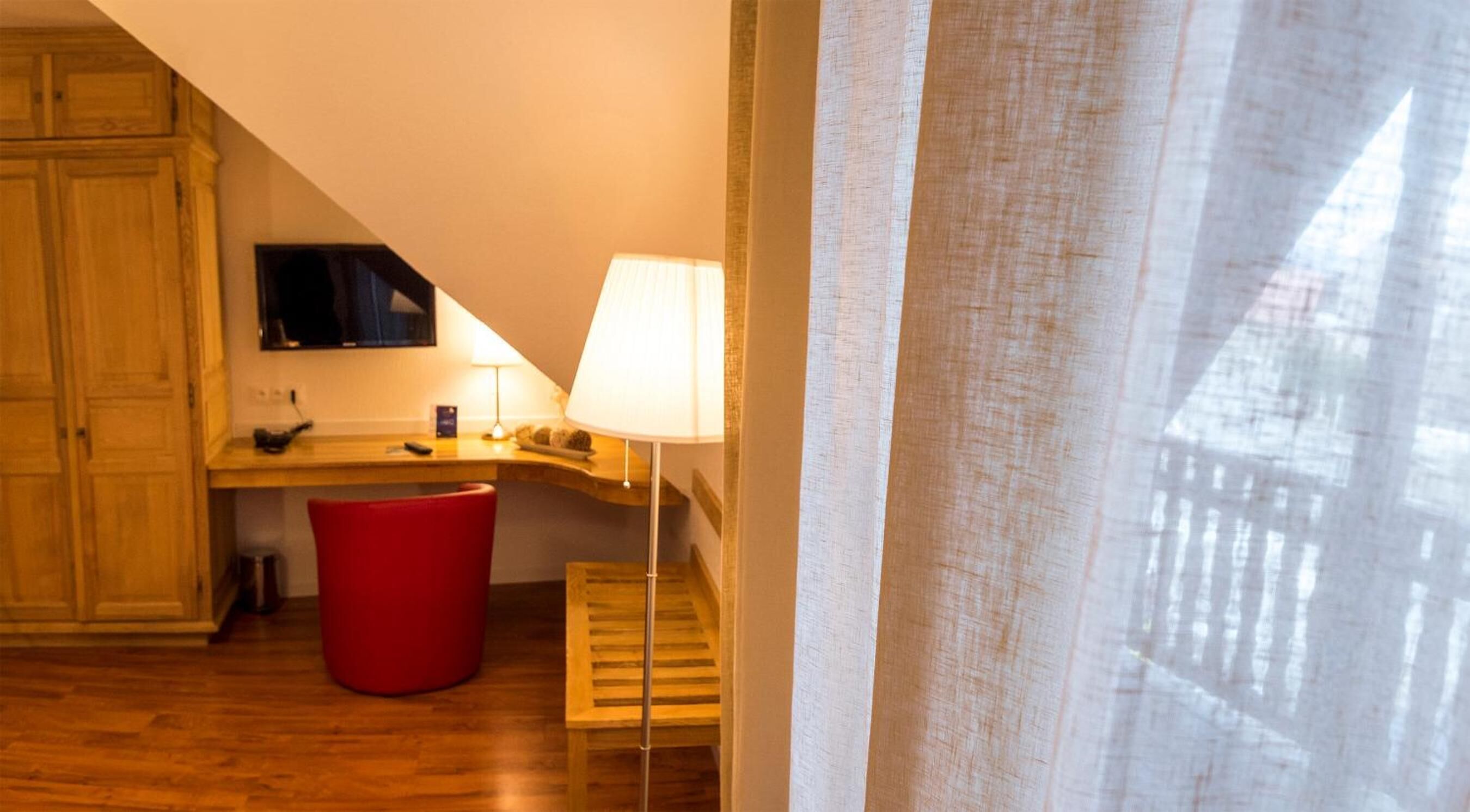Una habitación pequeña con un escritorio de madera, un televisor montado en la pared, una silla roja, una lámpara y piso de madera.