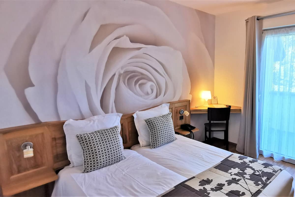 A hotel room with a large floral wall mural, a wooden headboard, two beds with white linens, patterned pillows, a desk with a lamp, and a view of a balcony with curtains.