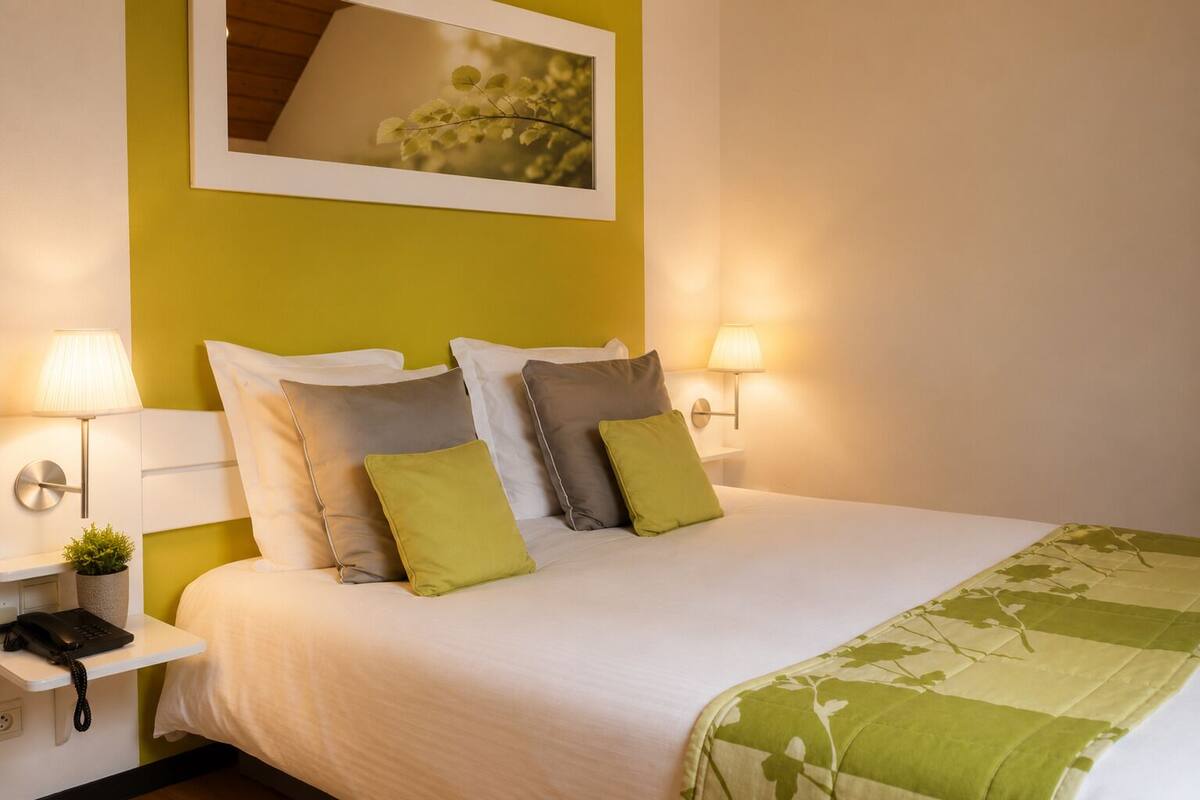 A neatly made bed with white and green bedding, two bedside lamps, a small plant, and a framed picture above the bed.