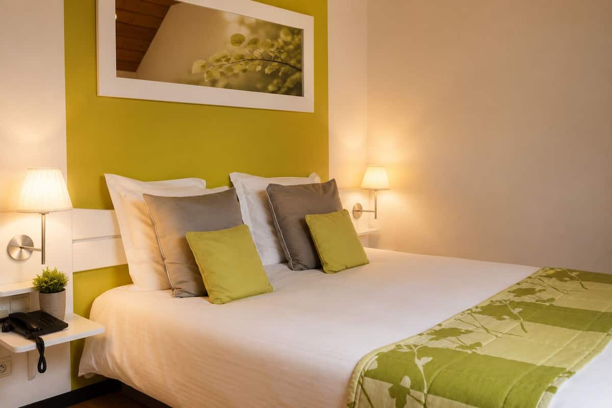 Una cama bien hecha con ropa de cama blanca y verde, dos lámparas de noche, una pequeña planta y un cuadro enmarcado sobre la cama.