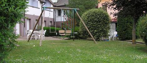 A grassy outdoor area with a swing set, white plastic chairs, and a bench.