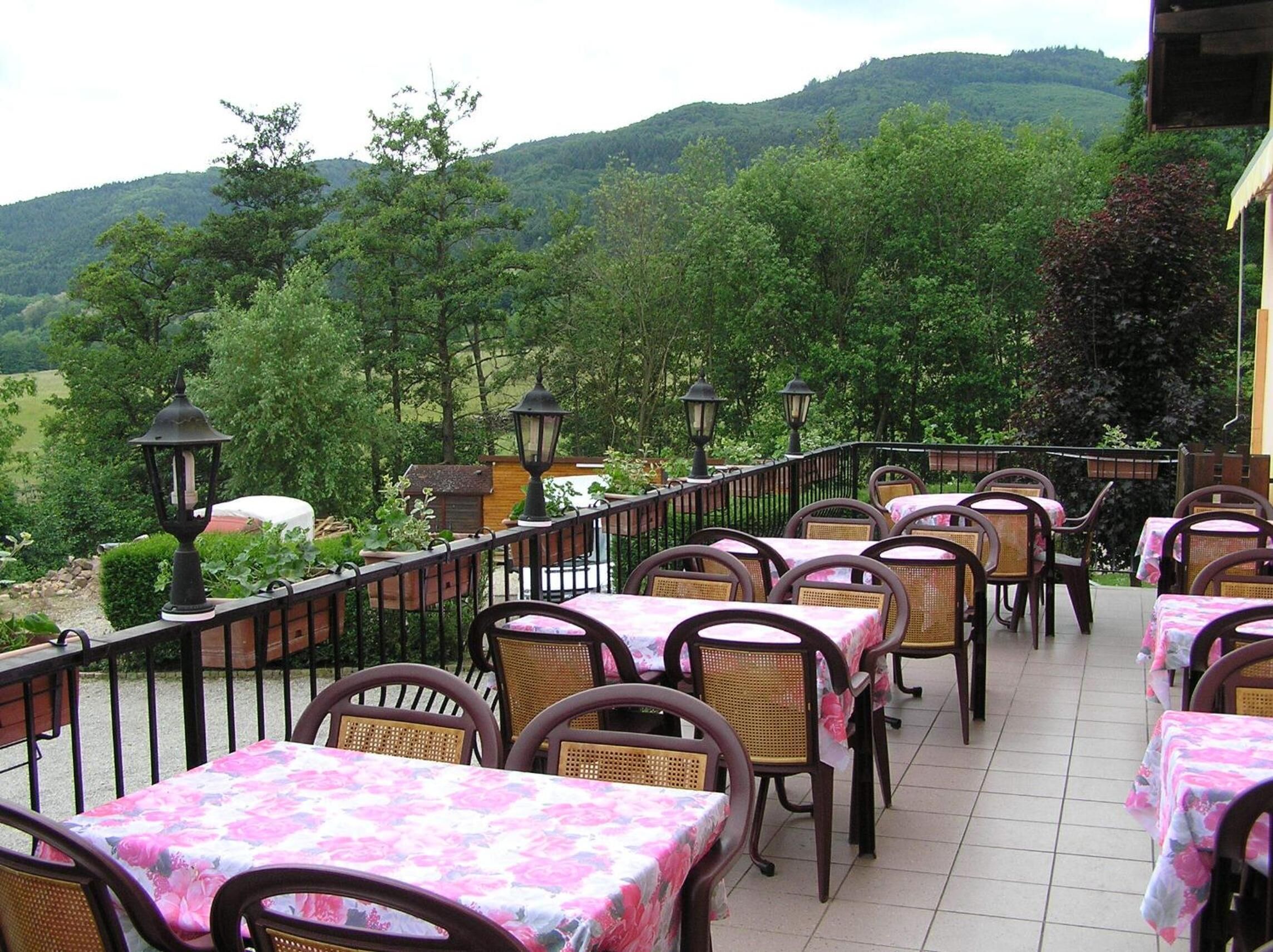 An outdoor patio with tables and chairs, surrounded by a metal railing, overlooking a lush green landscape with hills in the background.