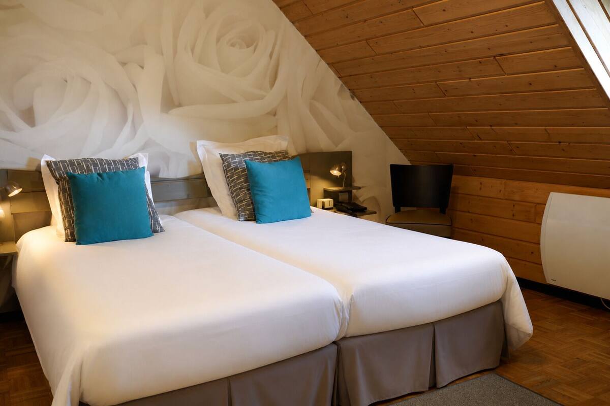 A room with two beds, a wooden ceiling, a wall with a floral design, and a bedside table with a lamp.