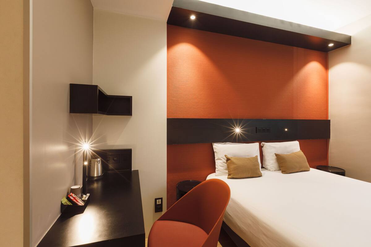 Ein modernes Hotelzimmer mit Bett, Nachttisch, einem roten Sessel und einem an der Wand befestigten Regal.