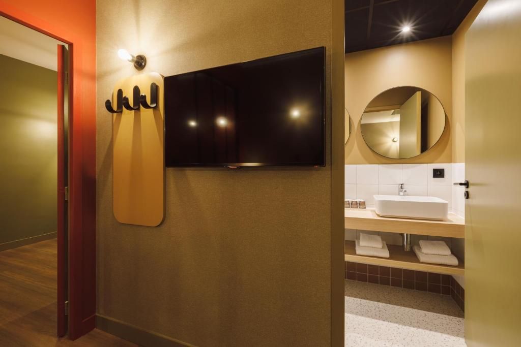 A modern bathroom with a flat-screen TV mounted on the wall, a round mirror, and a white sink with a wooden countertop.