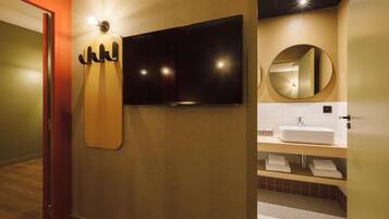 A modern bathroom with a flat-screen TV mounted on the wall, a round mirror, and a white sink with a wooden countertop.