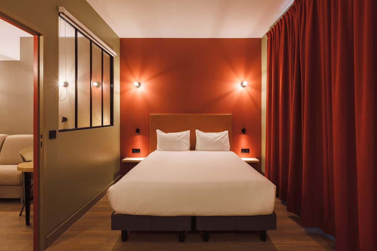 Ein Hotelzimmer mit einem großen Bett, zwei Nachttischen mit Lampen, einem roten Vorhang und einer Tür zu einem anderen Raum.