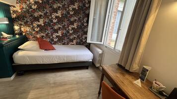 Une chambre avec un mur orné d’un motif floral, un lit avec une literie blanche et un oreiller rouge, un bureau en bois avec une chaise, et une fenêtre avec des rideaux.