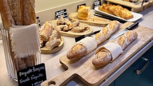Exposição de pães e doces numa padaria, incluindo baguetes e croissants, dispostos sobre tábuas de madeira e uma tábua de corte.