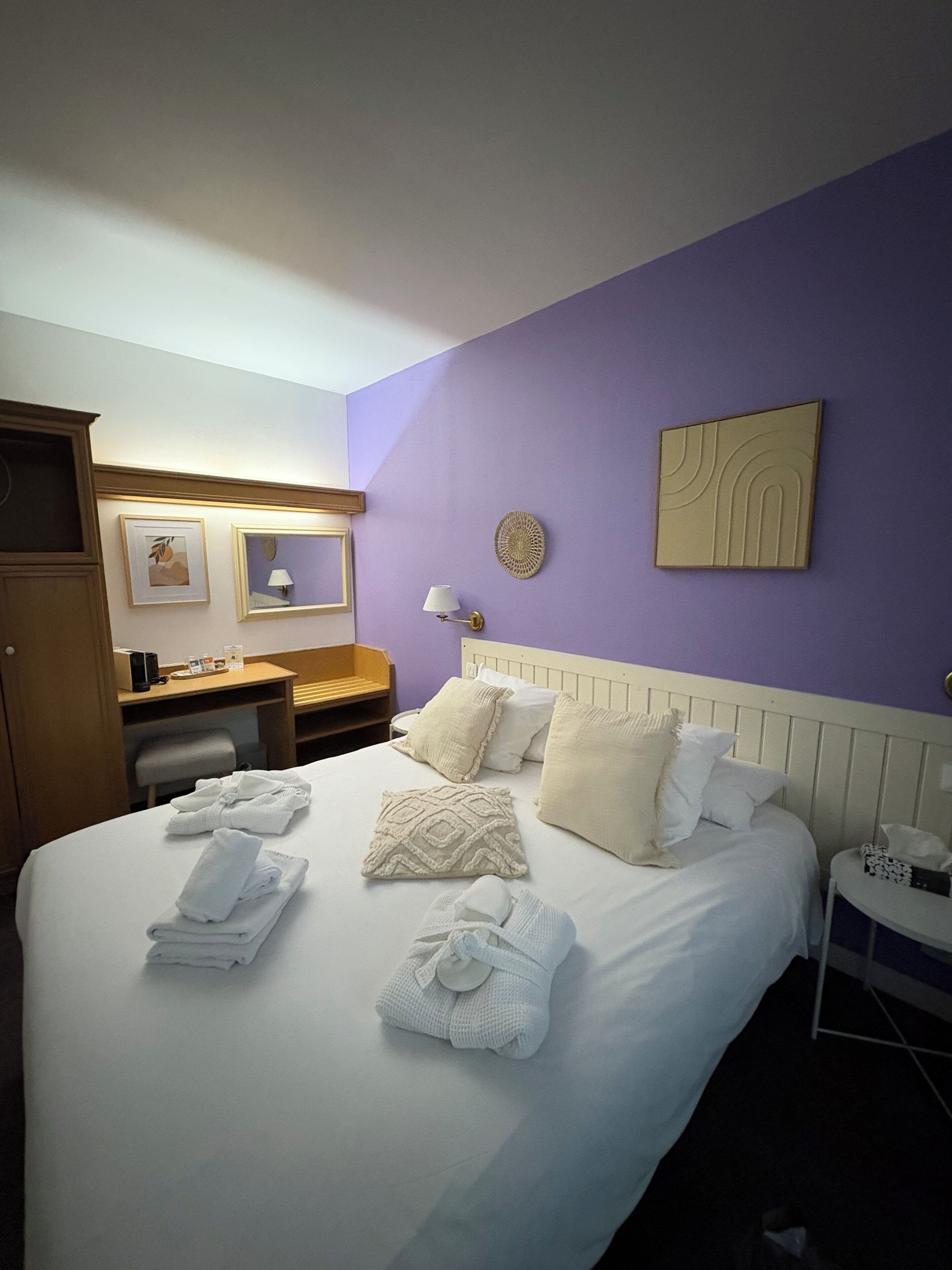 A hotel room with a large bed, a wooden wardrobe, a desk with a lamp, and a small table with a phone.