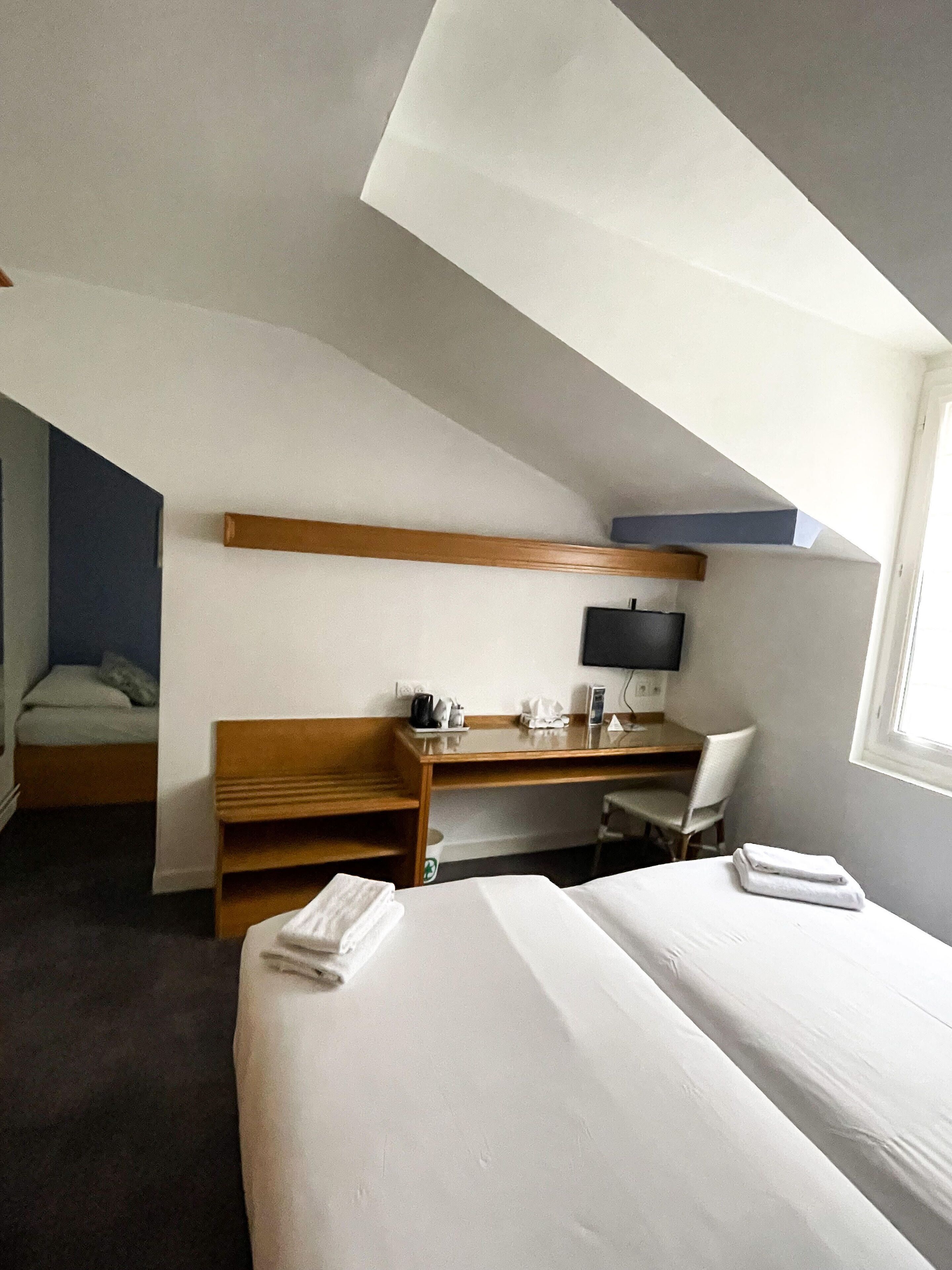 A hotel room with two beds, a desk, a chair, a TV, and a small window.