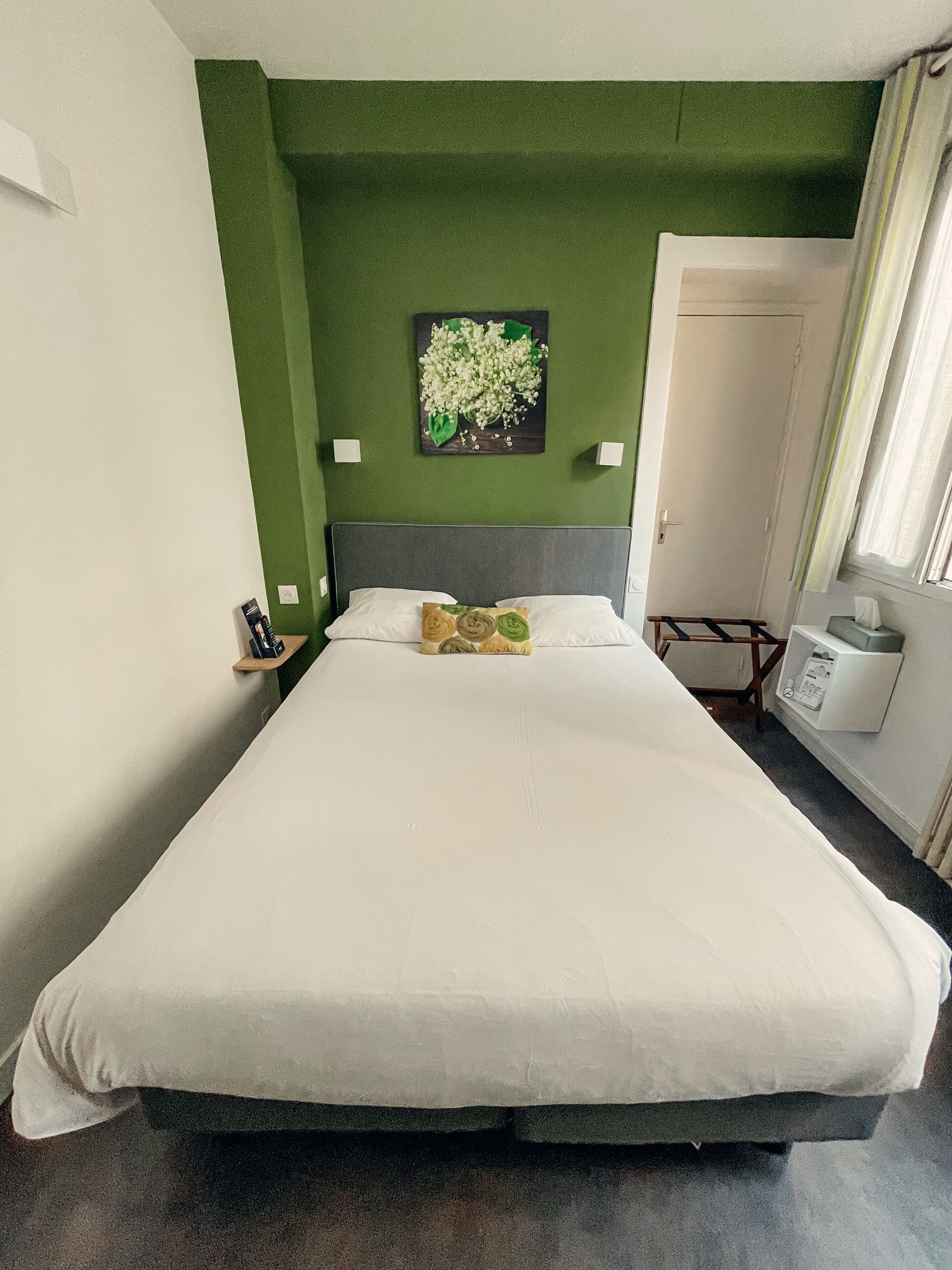 Ein Hotelzimmer mit Bett, Nachttisch, Wandbild und einem Fenster mit Vorhängen.
