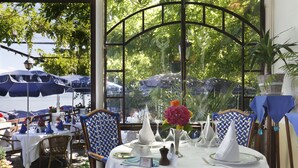 Una mesa dispuesta para una comida, con mantel blanco, sillas de estampado azul y blanco, y vista al mar a través de un ventanal.