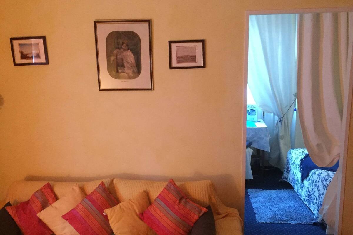 Sala de estar con un sofá, cuadros en la pared, una mesita con un control remoto y vistas a una habitación contigua a través de una cortina.