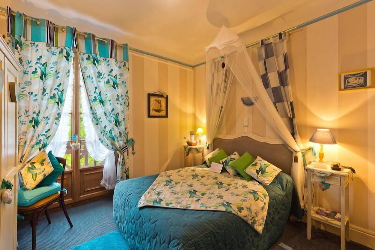 Un dormitorio con una cama, una silla, una mesita de noche, una lámpara, cortinas y una ventana.