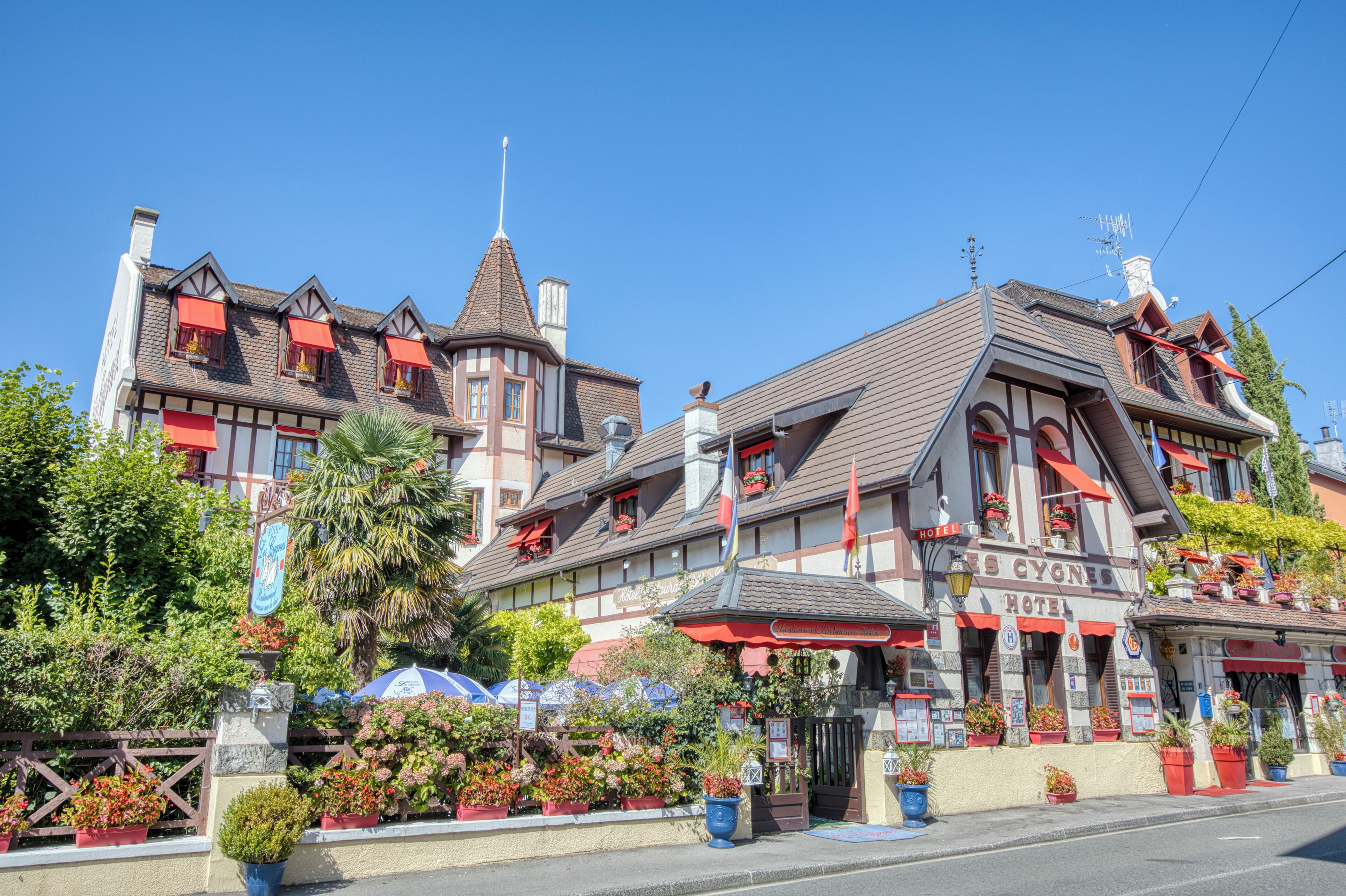 Un hôtel de style européen traditionnel, reconnaissable à son enseigne bien visible, à ses nombreuses fenêtres surmontées d’auvents et à la variété de plantes en pot qui ornent sa façade.