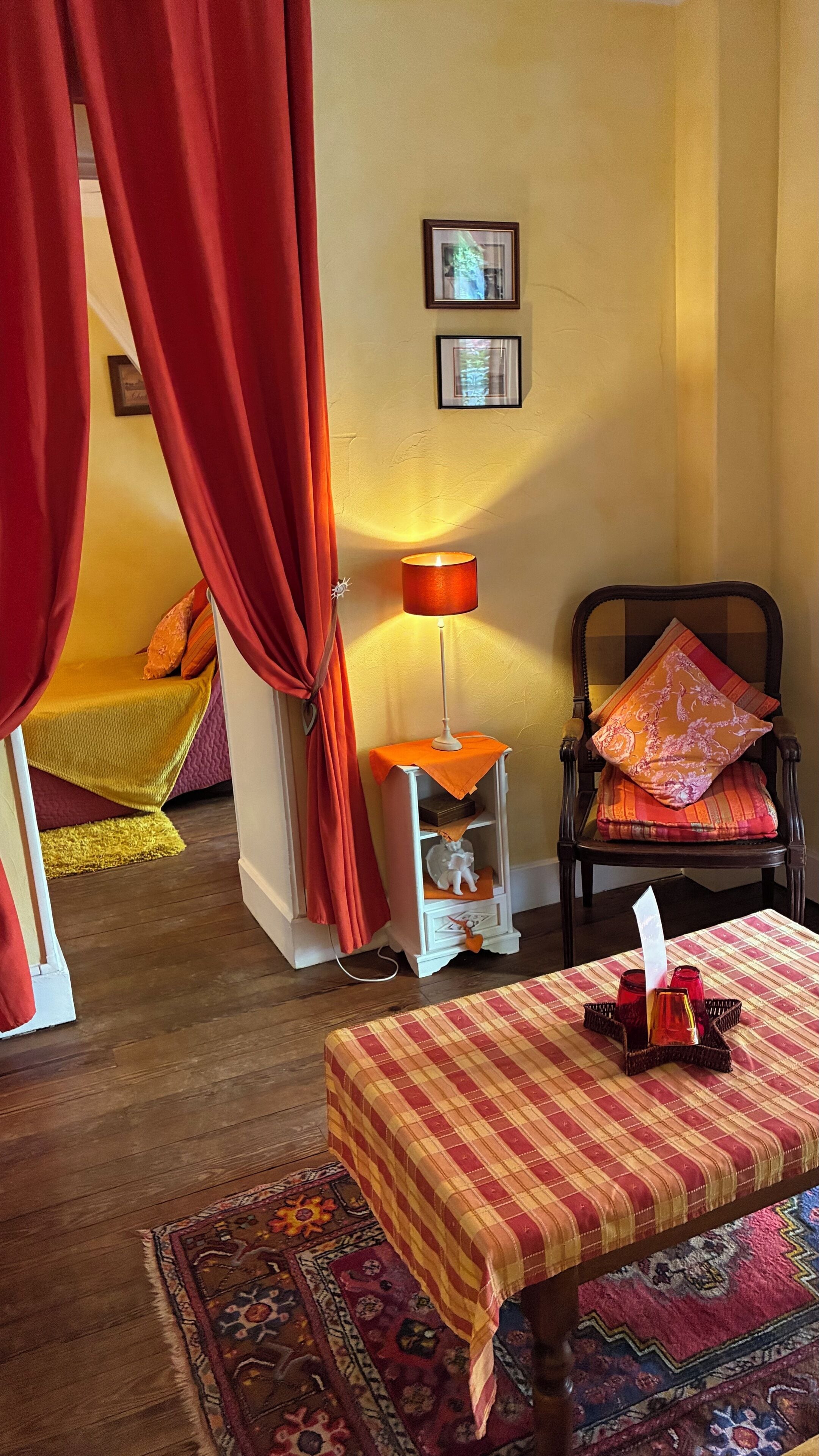 A cozy room with a bed, a table with candles, a chair with cushions, a lamp, and a rug.