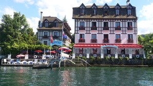 Un hotel tradicional europeo con un toldo rojo, una entrada visible y una masa de agua en primer plano.