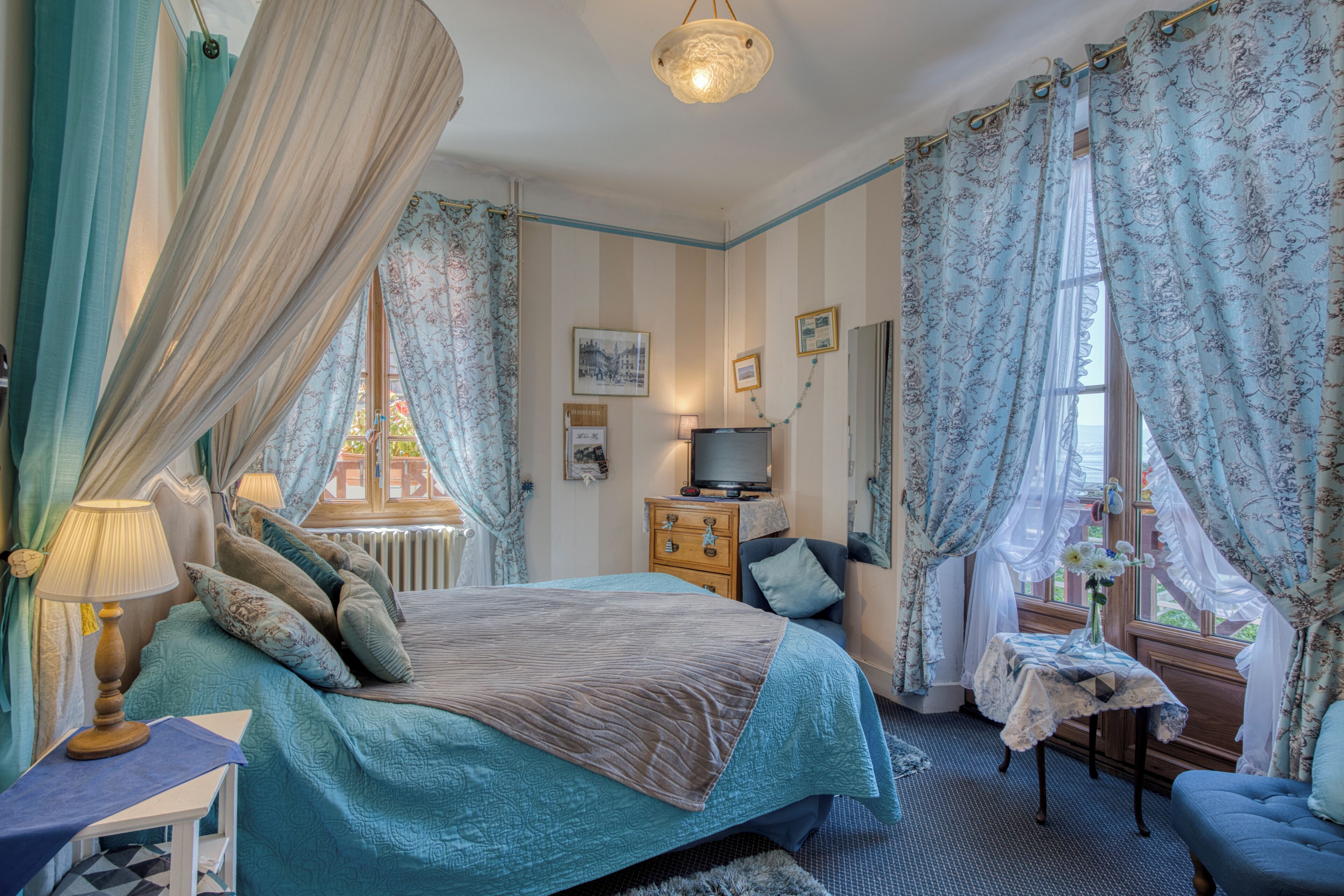 A bedroom with a bed, bedside table, lamp, dresser, TV, and curtains.