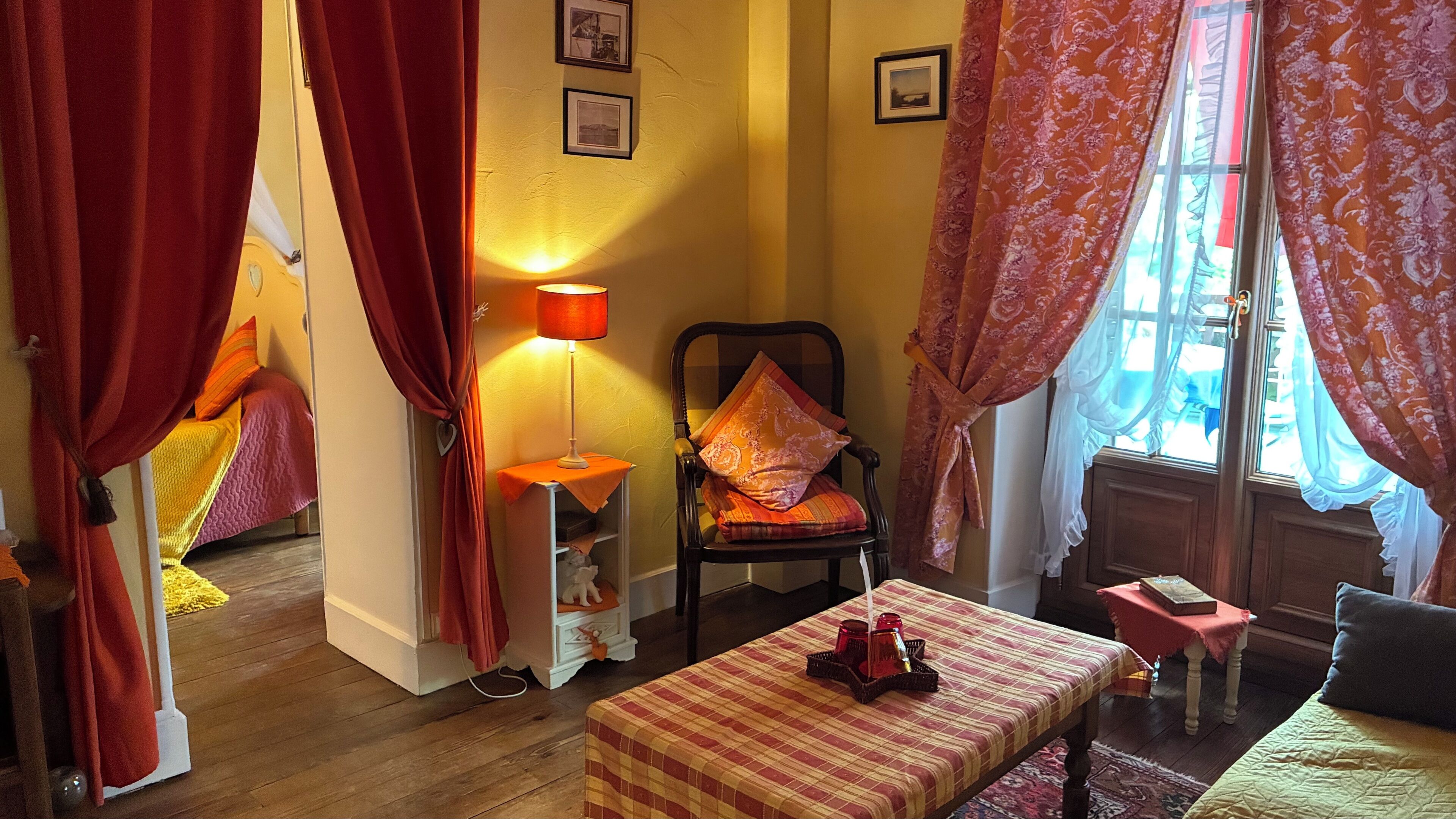 Una habitación acogedora con una cama, una silla con cojines, una mesa con lámpara y una ventana con cortinas.