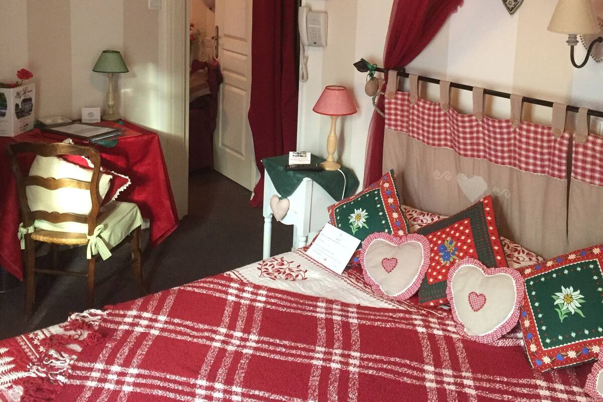 Un dormitorio con una colcha escocesa, una cama con dosel rojo, un escritorio con lámpara y una silla.