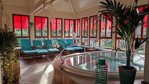 Un área de piscina interior iluminada por el sol, con suelo de madera, sillas azules acolchadas y una planta grande en maceta.