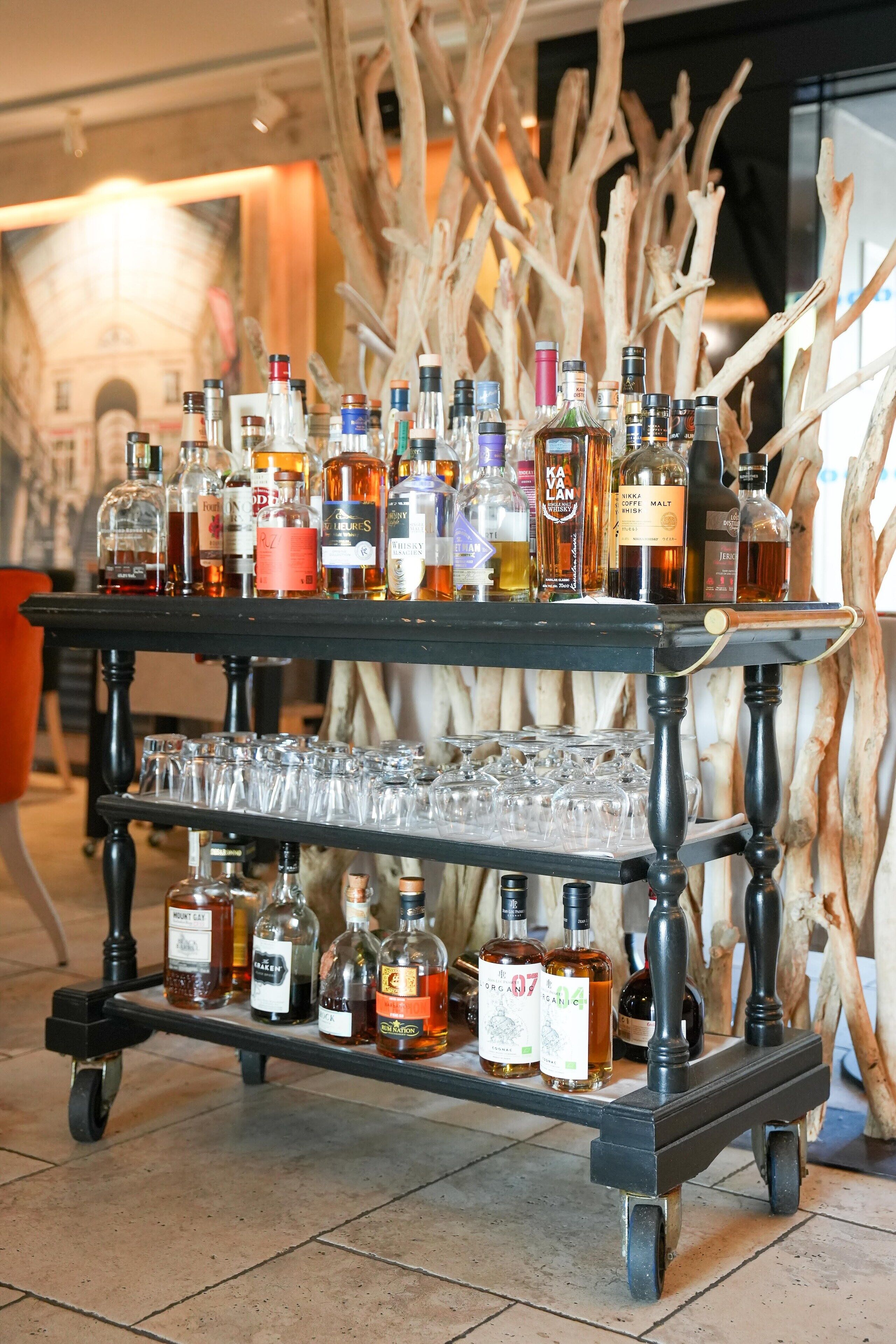 A bar cart with various bottles and glasses, set against a backdrop of wooden branches.