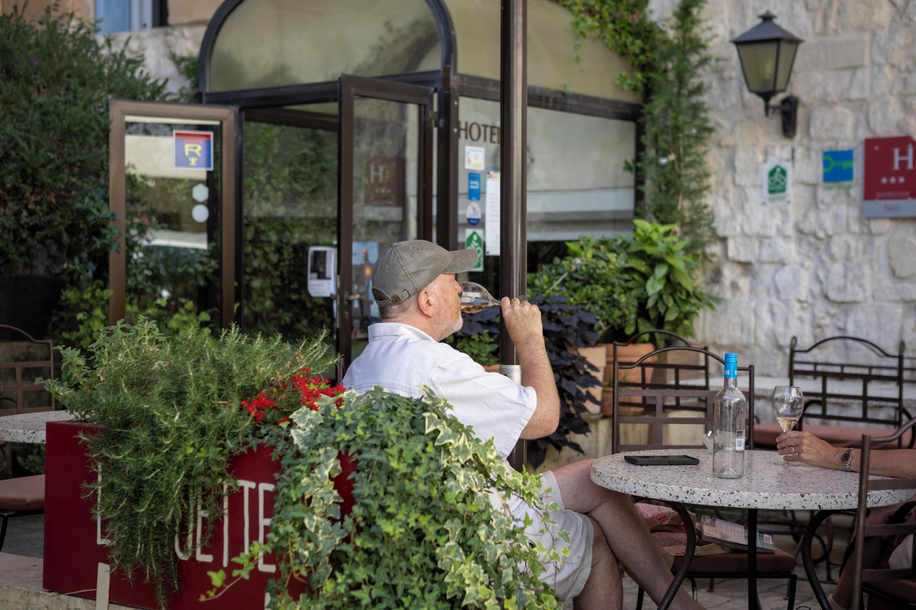 Un homme est assis à une table en plein air, devant une bouteille et un verre, un téléphone, et un panneau rouge sur lequel on peut lire le mot « HÔTEL ».
