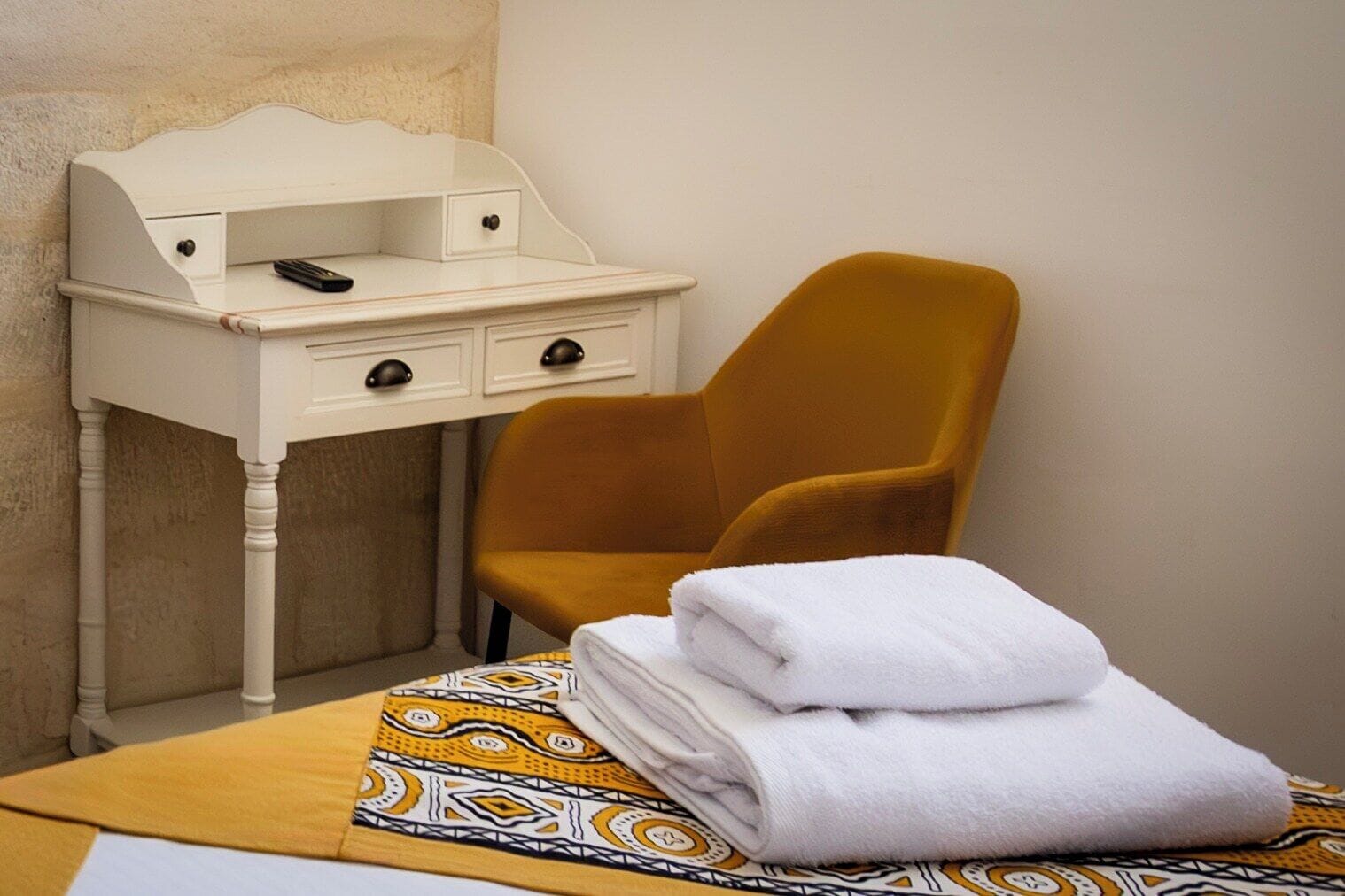 A hotel room with a bed, a white desk with drawers, a mustard-colored chair, and a neatly folded white towel on the bed.