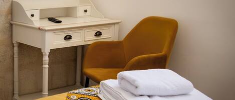 A hotel room with a bed, a white desk with drawers, a mustard-colored chair, and a neatly folded white towel on the bed.