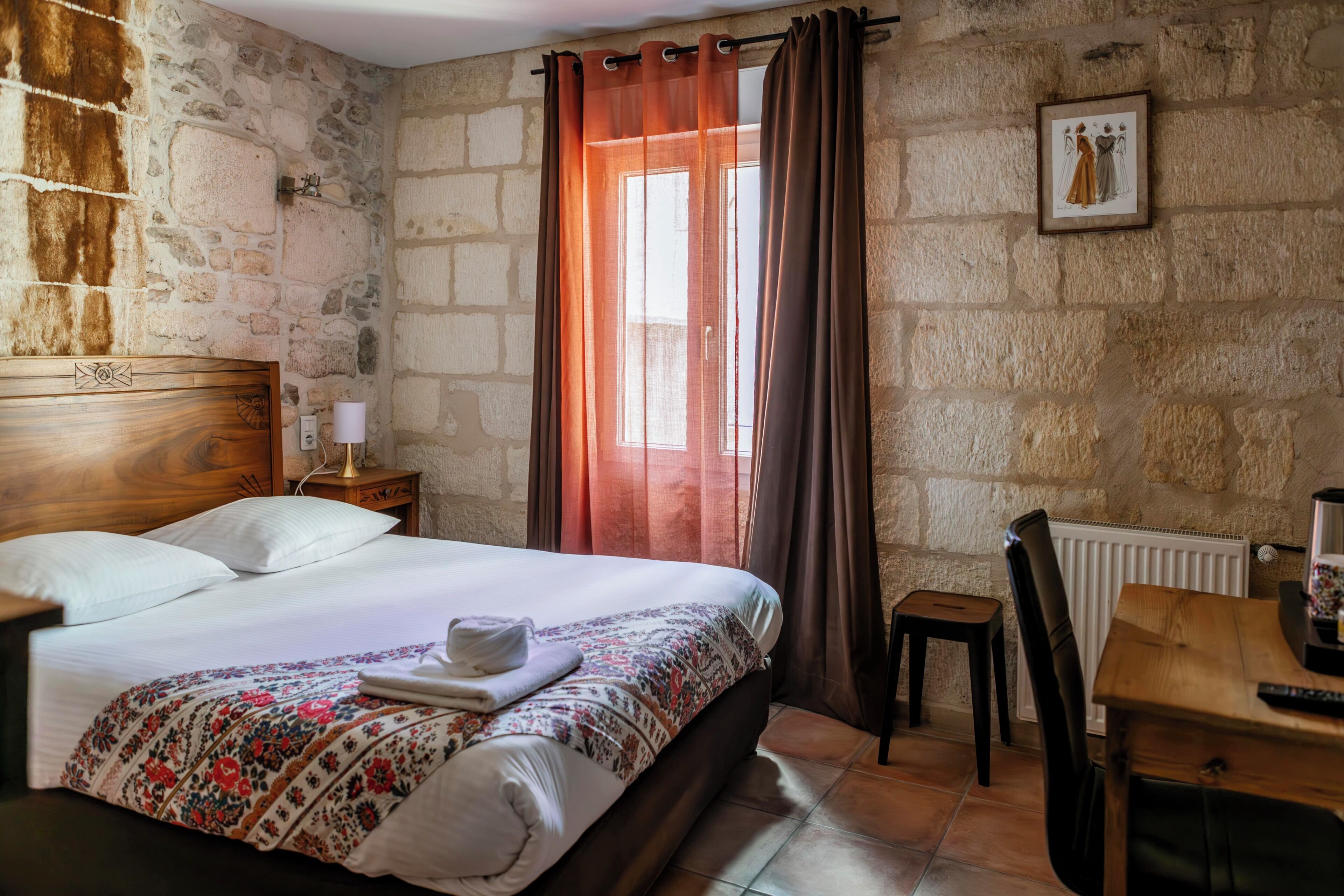 A bedroom with a stone wall, a wooden bed, a nightstand, a chair, a table, and a window with curtains.