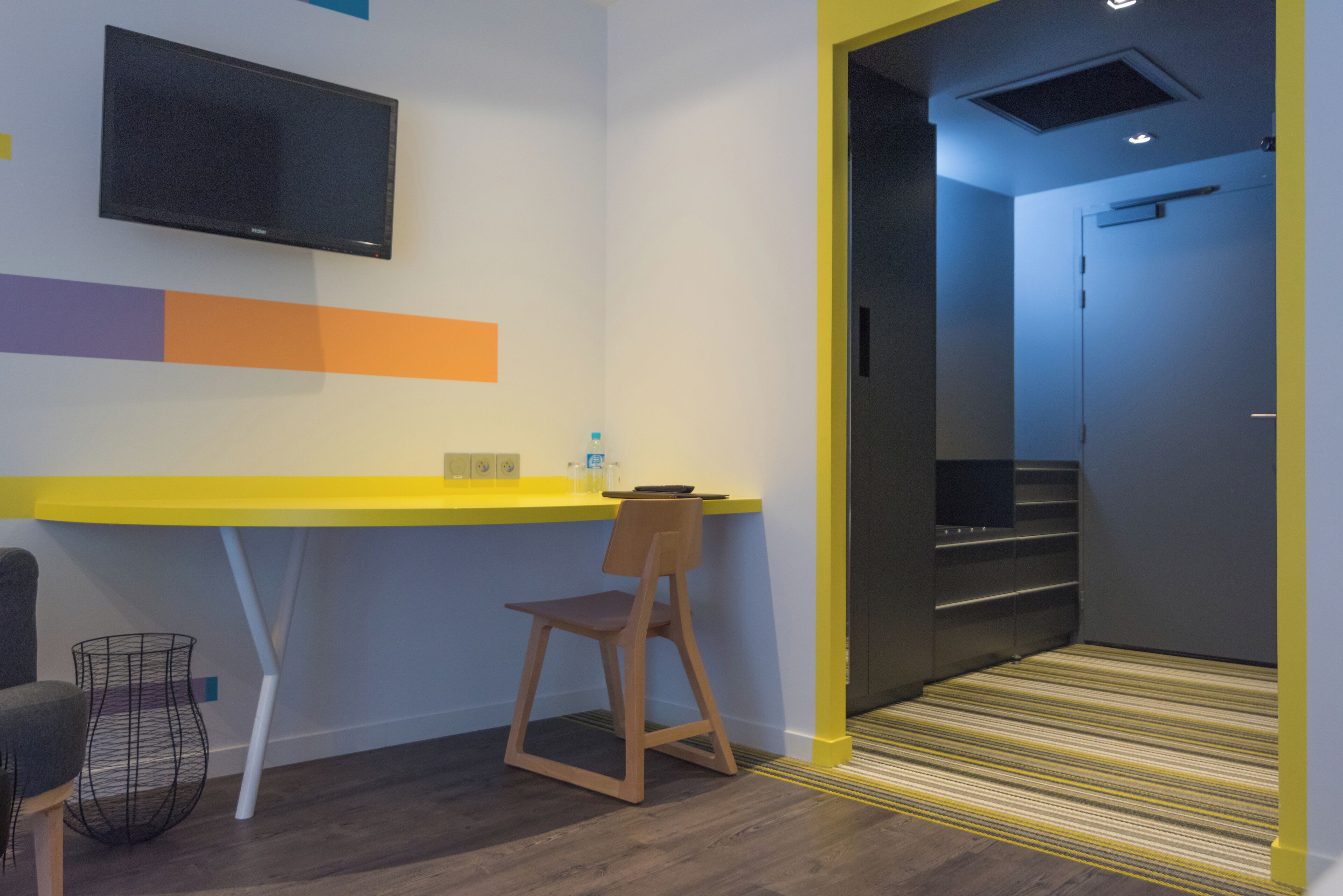 Een moderne kamer met een geel bureau, een stoel, een aan de muur bevestigde televisie en een deur die leidt naar een trap.